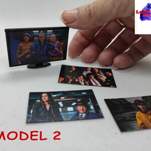 Op de afbeelding: Een miniatuurtelevisie met een zwart frame en een scherm waarop een groep mensen te zien is. Er liggen vier kleinere foto's van mensen op een wit oppervlak. De tekst "MODEL 2" is op het oppervlak gedrukt.