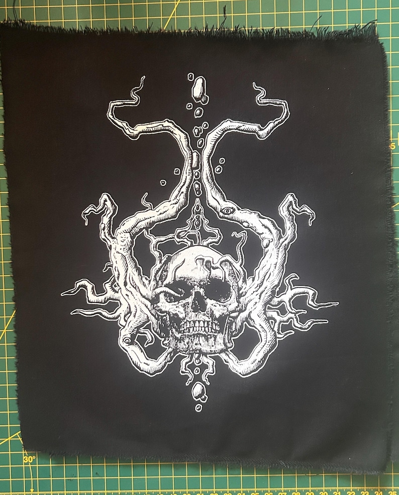 Op de afbeelding: Zwart vierkantige patch met een witte illustratie van een schedel met ingewikkelde, vertakte ontwerpen. De schedel heeft gedetailleerde tanden en is omgeven door organische, wervelende vormen. De randen van de patch zijn gerafeld.