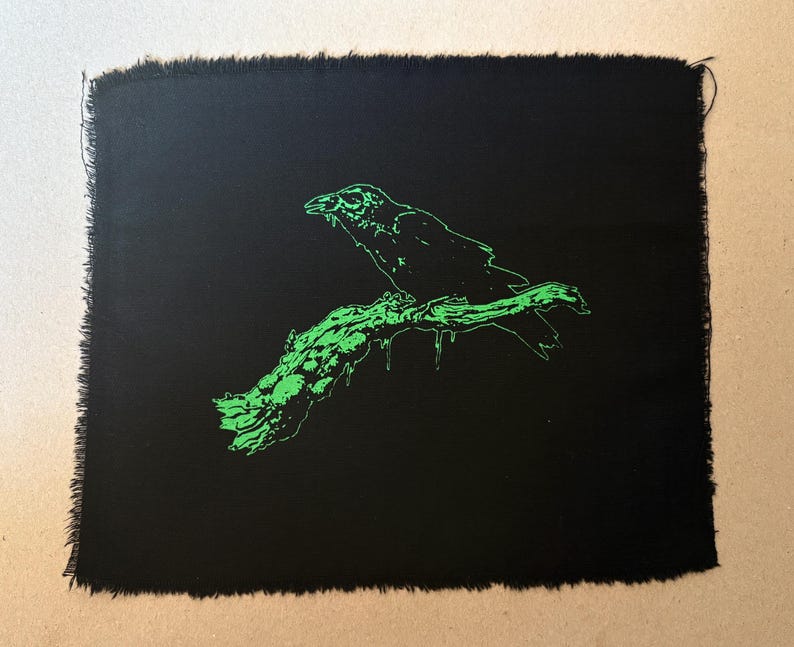 May include: Black fabric patch with a green illustration of a raven perched on a branch. The raven and branch are outlined in bright green against the black background. The edges of the patch are frayed.