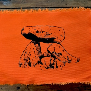 Puede incluir: Un parche rectangular de tela naranja con bordes deshilachados que presenta una ilustración en tinta negra de un dolmen prehistórico o estructura de tumba de piedra. El diseño muestra grandes rocas apiladas con líneas detalladas y hierba.