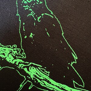 May include: Black fabric with a neon green outline of a crow perched on a branch. The bird is facing left, with its beak open. The branch is also outlined in neon green, creating a striking contrast against the dark background.