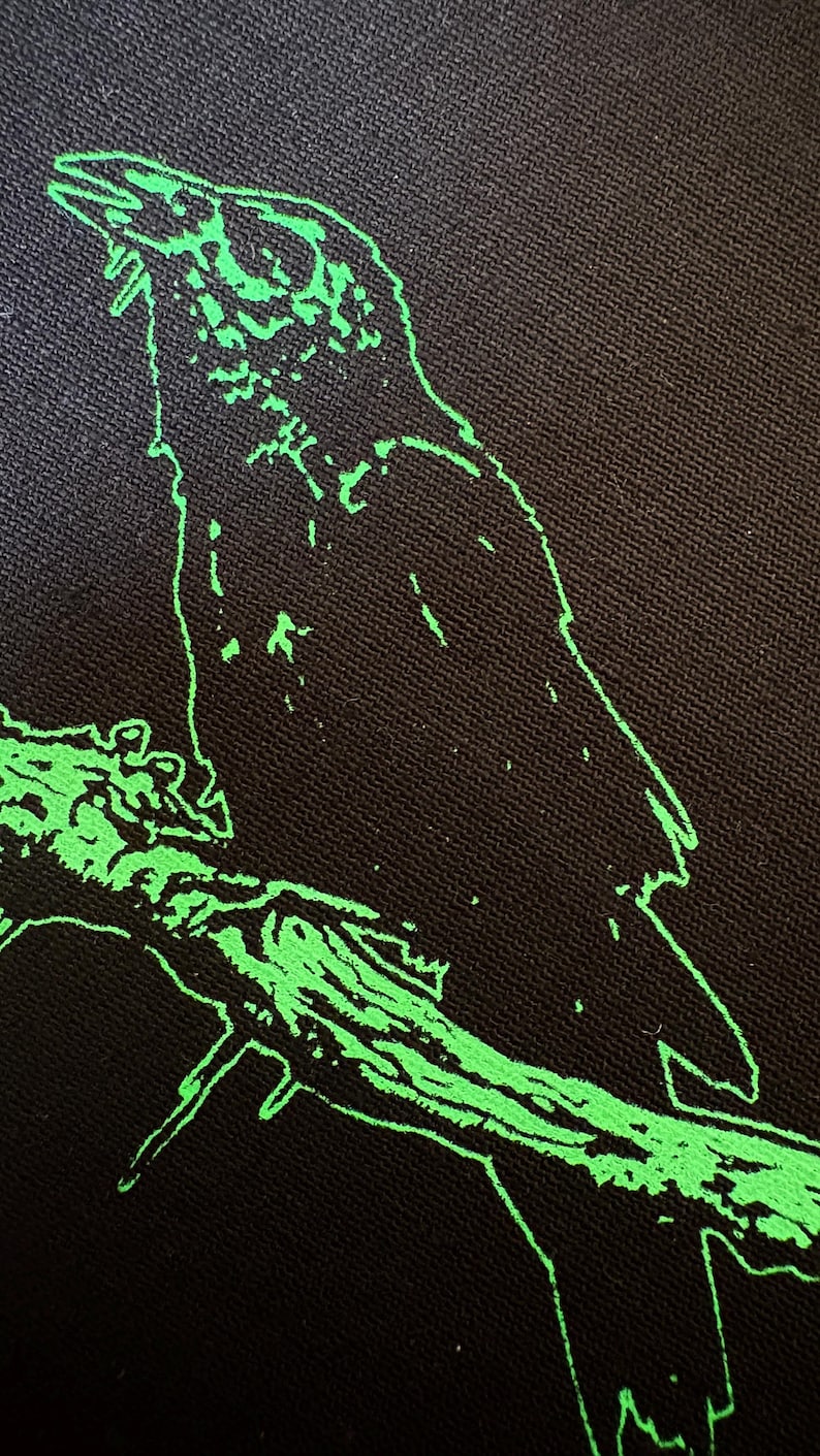 May include: Black fabric with a neon green outline of a crow perched on a branch. The bird is facing left, with its beak open. The branch is also outlined in neon green, creating a striking contrast against the dark background.