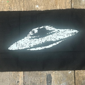 May include: A white UFO silhouette on a black fabric background.