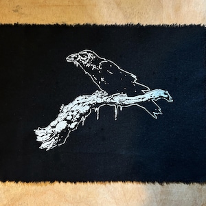 May include: A black rectangular fabric patch featuring a white illustration of a raven on a branch. The raven is depicted in profile, and the branch has a detailed texture. The patch's edges are intentionally frayed.