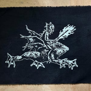 May include: Black patch with a white illustration of a horned figure riding a frog. The figure holds a flaming object. The patch has a raw, unfinished edge. The design is a bold, graphic style.