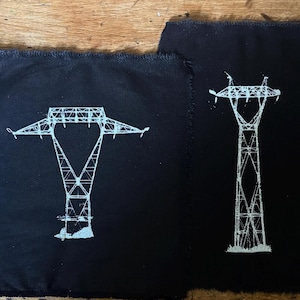 May include: Four rectangular black fabric patches with frayed edges, each featuring a white screen-printed illustration of a different style of electrical transmission tower. The designs display intricate lattice structures of various pylon shapes.