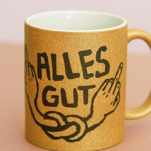Könnte beinhalten: Goldener Glitzertasse mit den schwarzen Worten "ALLES GUT" und einer handgezeichneten Illustration von zwei Händen, die den Mittelfinger zeigen. Die Tasse hat einen weißen Innenraum und einen goldenen Griff.