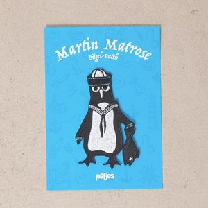 Könnte beinhalten: Ein schwarz-weißer Pinguin-Aufnäher mit Matrosenmütze und -uniform, der einen Fisch hält. Der Aufnäher befindet sich auf einer blauen Karte mit dem Text "Martin Matrose Bügel Patch" und der Marke "pafjes".