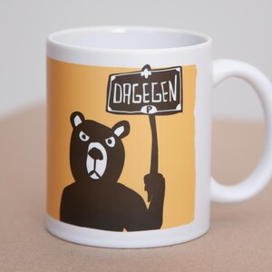 Könnte beinhalten: Weiße Keramiktasse mit einer Illustration eines schwarzen Bären, der ein Schild mit dem Wort "DAGEGEN" hält. Der Hintergrund ist orange. Die Tasse hat einen weißen Henkel und ist für Heißgetränke geeignet.