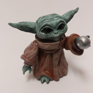 May include: A small green and brown figurine of a character from the Star Wars franchise, holding a silver sphere in its right hand.
