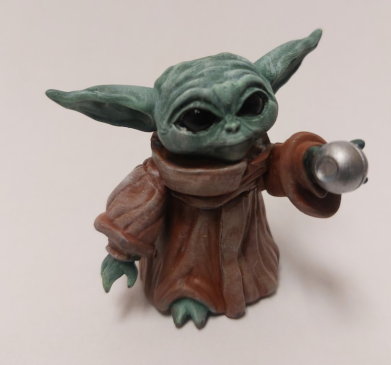 May include: A small green and brown figurine of a character from the Star Wars franchise, holding a silver sphere in its right hand.