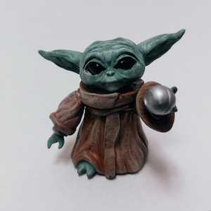 May include: A small green and brown figurine of a character from the Star Wars franchise, holding a silver sphere in its right hand.