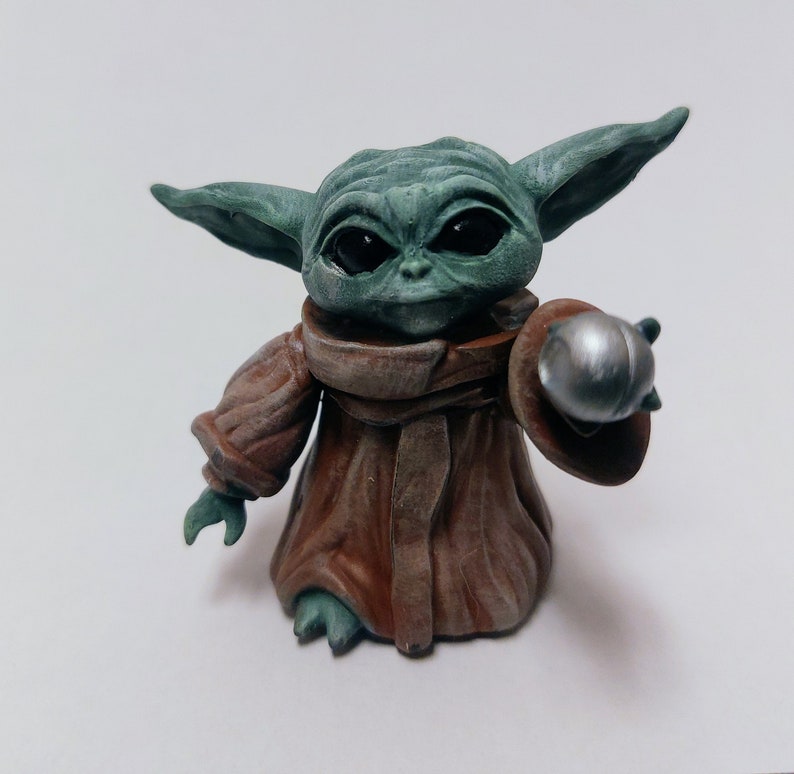 May include: A small green and brown figurine of a character from the Star Wars franchise, holding a silver sphere in its right hand.