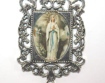 May include: Silver-toned metal frame with an oval-shaped image of the Virgin Mary in a blue robe. The image is surrounded by a white border and a silver-toned frame. The frame has a decorative design with floral motifs.