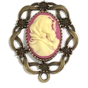 May include: A cameo pendant featuring a white cameo of a woman holding a child, set in a bronze-colored frame with a pink background. The frame has a decorative design with floral motifs.