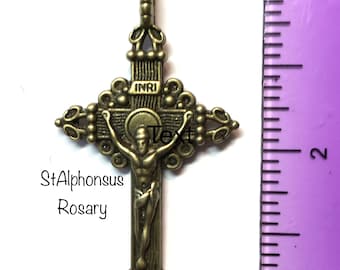 May include: Bronze-colored crucifix pendant with ornate detailing. The cross features a figure of Jesus Christ and the letters INRI. The text "StAlphonsus Rosary" is also visible. The pendant is shown next to a ruler.