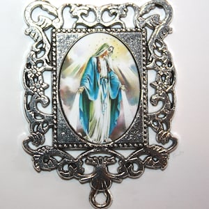 May include: Silver-toned metal pendant featuring a framed image of the Virgin Mary in a blue robe with a white veil. The pendant has a decorative filigree design and a loop for attaching to a chain.
