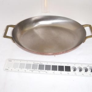 May include: A copper-bottomed, oval-shaped pan with a stainless steel interior. The pan has two brass handles. A color calibration chart and ruler are in the foreground.