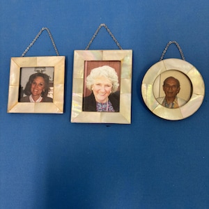 May include: Three small, decorative picture frames with mother-of-pearl borders. Two are square, and one is round. Each frame holds a photograph and hangs from a silver chain. The frames are displayed against a blue background.
