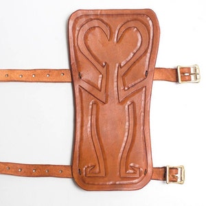 Puede incluir: Una espinillera de cuero marr&oacute;n con un dise&ntilde;o decorativo y dos correas con hebillas.