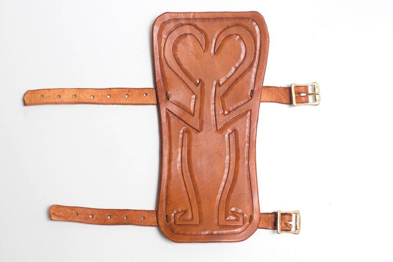 Puede incluir: Una espinillera de cuero marr&oacute;n con un dise&ntilde;o decorativo y dos correas con hebillas.