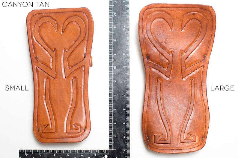 Puede incluir: Dos protectores de brazo de cuero, uno peque&ntilde;o y otro grande, en color marr&oacute;n ca&ntilde;&oacute;n. Ambos protectores tienen un dise&ntilde;o en forma de coraz&oacute;n con detalles intrincados. Los protectores est&aacute;n colocados planos sobre una superficie blanca con una regla para la escala.