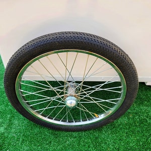 May include: A close-up of a black rubber tire with a silver spoke wheel. The tire is on a green artificial grass surface.