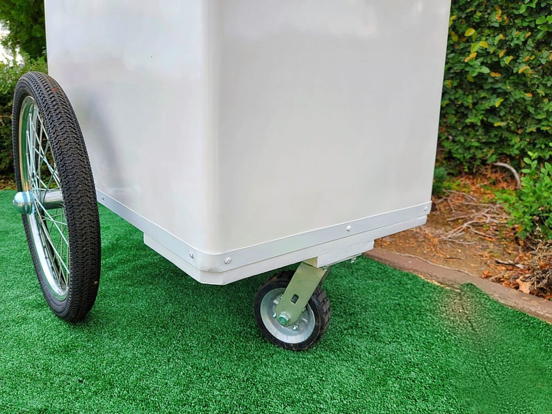 May include: White utility cart with a large rectangular body and four wheels. The cart has a silver trim around the edges of the body and the wheels are silver with black tires.