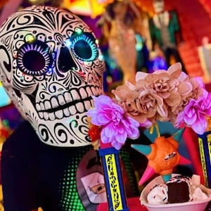 May include: A decorative skull mask with intricate black and white patterns, illuminated eyes, and a full set of teeth. The mask is held with a colorful arrangement of flowers, a star-shaped object, and a small bowl of dessert, creating a festive atmosphere.