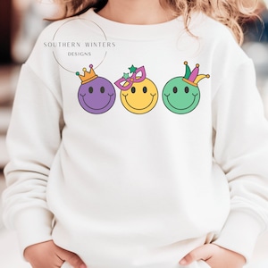 May include: White sweatshirt with a purple, yellow, and green smiley face design. Each smiley face has a different Mardi Gras themed accessory. The text "SOUTHERN WINTERS DESIGNS" is printed above the smiley faces.