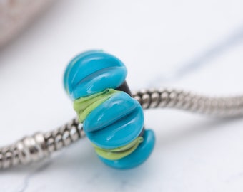 May include: A glass charm bead featuring a vibrant turquoise blue and lime green striped pattern, threaded onto a silver-toned snake chain bracelet. The bead has a rounded, ribbed texture with a glossy finish and a dark core visible at the center.
