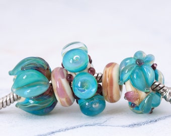 May include: A silver snake chain bracelet featuring several glass beads in shades of turquoise, teal, and soft pink. The beads include floral-shaped designs, smooth rounded shapes, and small dark purple accents, all strung together on a metal chain.