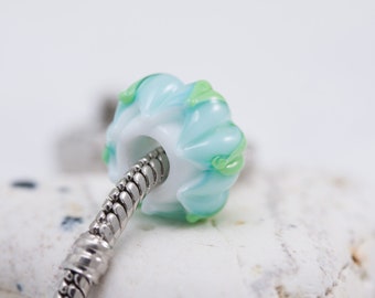 May include: A round glass bead with a white core and a light blue and lime green swirled pattern. The bead is threaded onto a silver-toned metal snake chain bracelet, featuring a textured, flexible design with a polished metallic finish.