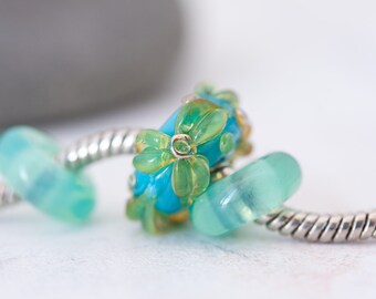 May include: A silver charm bracelet chain featuring three glass beads. The central bead is bright blue with raised, translucent light green floral patterns. Flanking it are two smooth, translucent light turquoise beads with a soft, glossy finish.