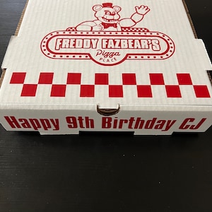 Może przedstawiać: Białe pudełko na pizzę z czerwono-białym wzorem w kratkę. Pudełko ma logo "Freddy Fazbear's Pizza Place" z kresk&oacute;wkowym niedźwiedziem w muszce. Tekst "Happy 9th Birthday CJ" jest nadrukowany na spodzie pudełka.