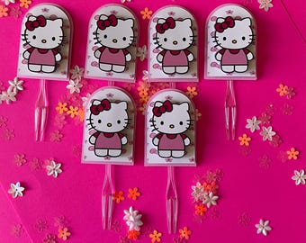 May include: Seven Hello Kitty cake toppers on a bright pink background, decorated with small flower accents. Each topper features the iconic cartoon cat in a pink dress and bow. The toppers have clear plastic sticks.