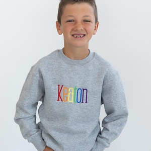 May include: A gray sweatshirt with the name "Keaton" embroidered in rainbow colors. The sweatshirt is a crew neck style and is worn by a person. The person is wearing blue jeans.