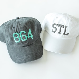 May include: Two baseball caps, one black with the number "864" embroidered in teal green, and one white with "STL" embroidered in black.