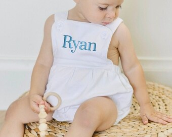 May include: A white baby romper with blue embroidered text that says "Ryan". The romper has a gathered skirt and is worn by a baby sitting on a woven straw cushion.
