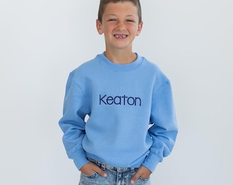 Puede incluir: Un niño pequeño con una sudadera azul claro con el nombre "Keaton" bordado en la parte delantera. También lleva pantalones vaqueros azules y calcetines grises.