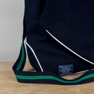 Puede incluir: Una bolsa de lona azul marino con ribete blanco y una correa verde y negra. La bolsa tiene una etiqueta que dice "ShoreBags" y "Materiales sostenibles Lona natural Eco-positiva Reciclable/Biodegradable".