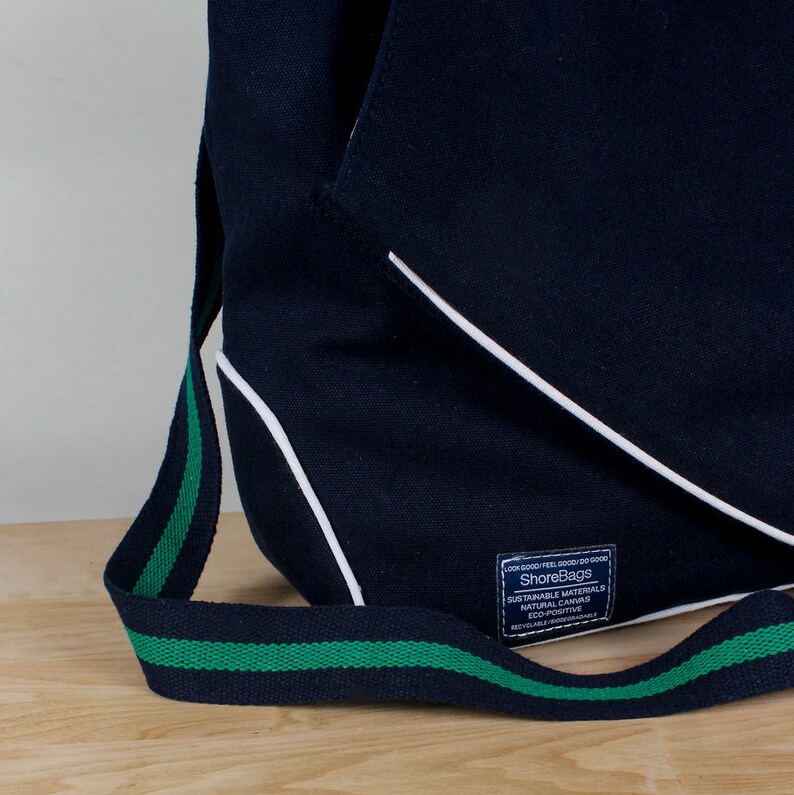 Puede incluir: Una bolsa de lona azul marino con ribete blanco y una correa verde y negra. La bolsa tiene una etiqueta que dice "ShoreBags" y "Materiales sostenibles Lona natural Eco-positiva Reciclable/Biodegradable".