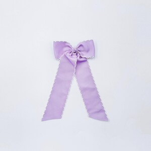 May include: A light purple hair bow made of ribbed fabric with a scalloped white edge. The bow features a central knot and two long, hanging tails that extend downwards, showcasing a consistent texture and a clean, symmetrical design throughout.