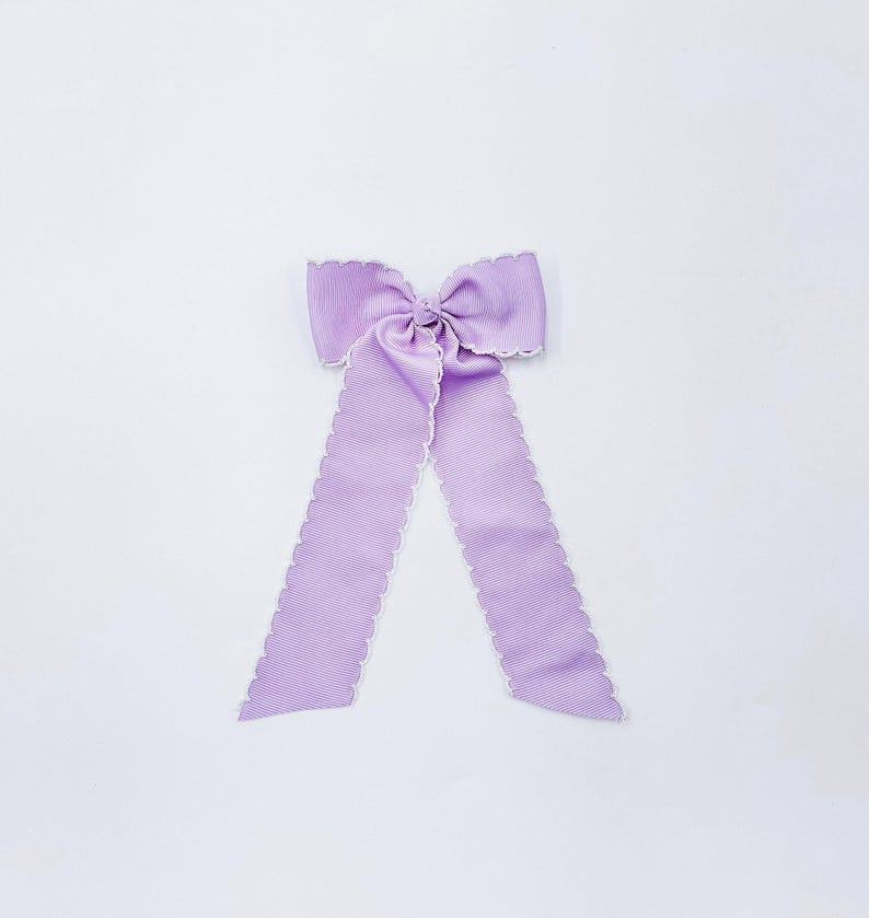 May include: A light purple hair bow made of ribbed fabric with a scalloped white edge. The bow features a central knot and two long, hanging tails that extend downwards, showcasing a consistent texture and a clean, symmetrical design throughout.