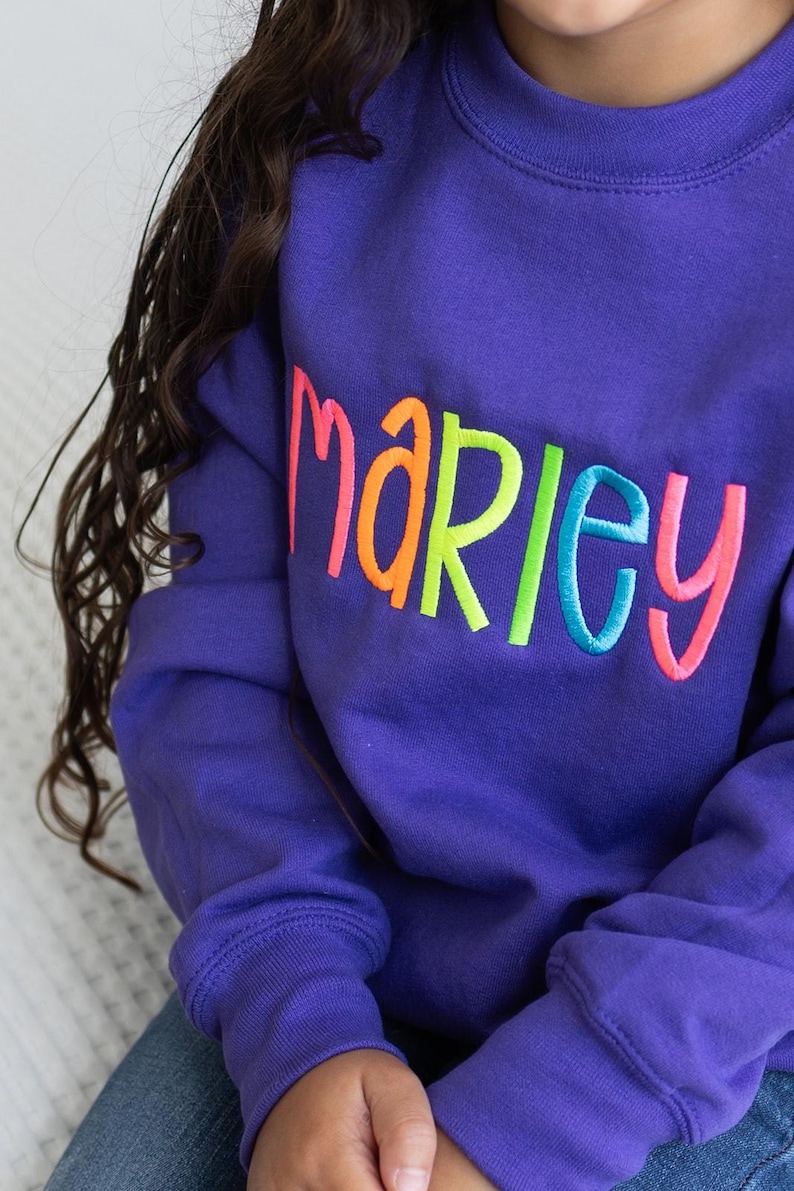 Puede incluir: Sudadera morada con el nombre "Marley" bordado en colores arco&iacute;ris.