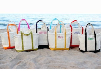 May include: A collection of personalized canvas tote bags with colorful accents. Each bag features a different name embroidered in a coordinating color, with handles and trim in various hues like orange, pink, green, blue, yellow, red, and black. The bags are displayed on a sandy beach.