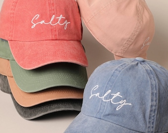 May include: A stack of six baseball caps in various colors, including red, green, brown, gray, and blue. Each cap has the word "Salty" embroidered on the front in white.