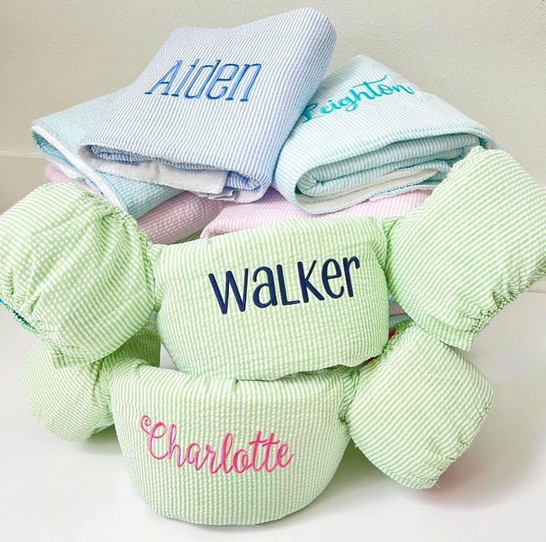 May include: A stack of green and white striped towels with the names "Walker", "Charlotte", and "Aiden" embroidered on them. There is also a light blue and white striped towel with the name "Feighton" embroidered on it.