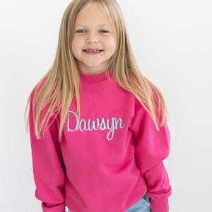 Puede incluir: Una sudadera rosa con el nombre "Dawson" bordado en azul claro en la parte delantera. La sudadera la lleva puesta una persona joven con el pelo largo y rubio.