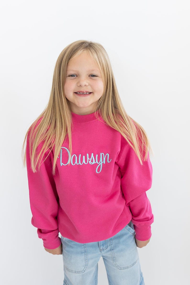 Puede incluir: Una sudadera rosa con el nombre "Dawson" bordado en azul claro en la parte delantera. La sudadera la lleva puesta una persona joven con el pelo largo y rubio.